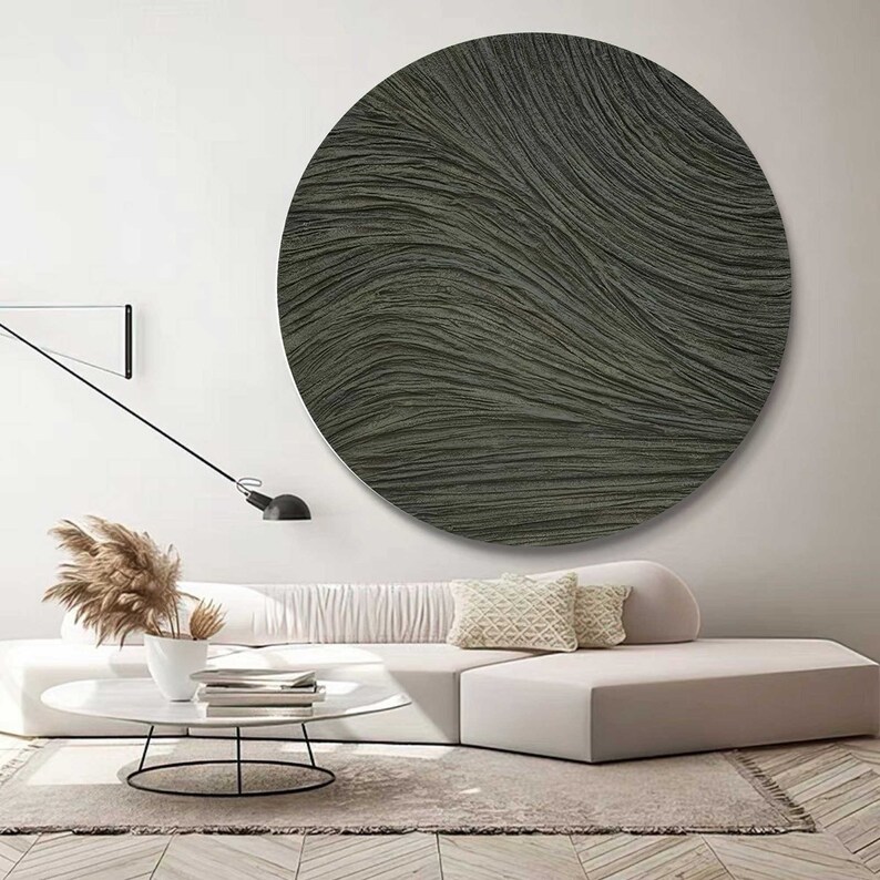 May include: A large, round, textured wall art piece in shades of gray and olive green hangs above a white sofa. The artwork features a rippled, wave-like pattern. The room is decorated in neutral tones with a modern aesthetic.