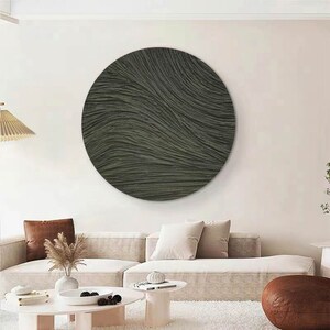 May include: A large, circular wall art piece with a textured, wave-like pattern in shades of gray and green. The artwork is mounted on a white wall above a beige sofa, creating a modern and minimalist aesthetic. The room is decorated with neutral tones.