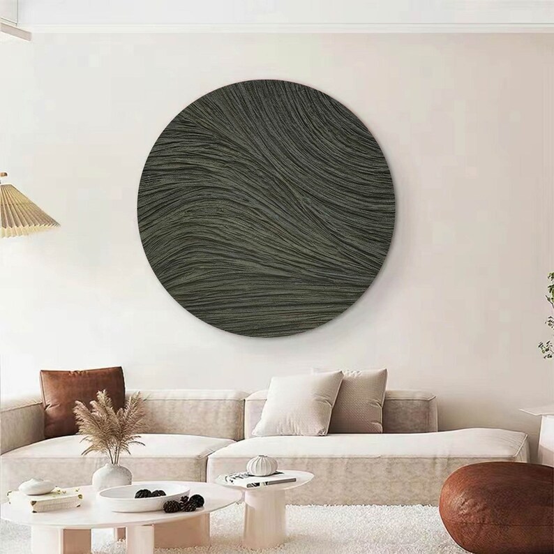 May include: A large, circular wall art piece with a textured, wave-like pattern in shades of gray and green. The artwork is mounted on a white wall above a beige sofa, creating a modern and minimalist aesthetic. The room is decorated with neutral tones.