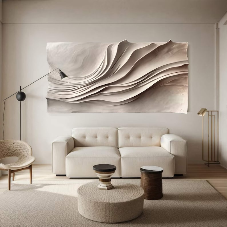May include: A neutral-toned living room featuring a large, abstract wall art piece with layered, wave-like forms. A white sofa, a round woven ottoman, and two small side tables complete the scene. A black floor lamp and a gold floor lamp provide lighting.