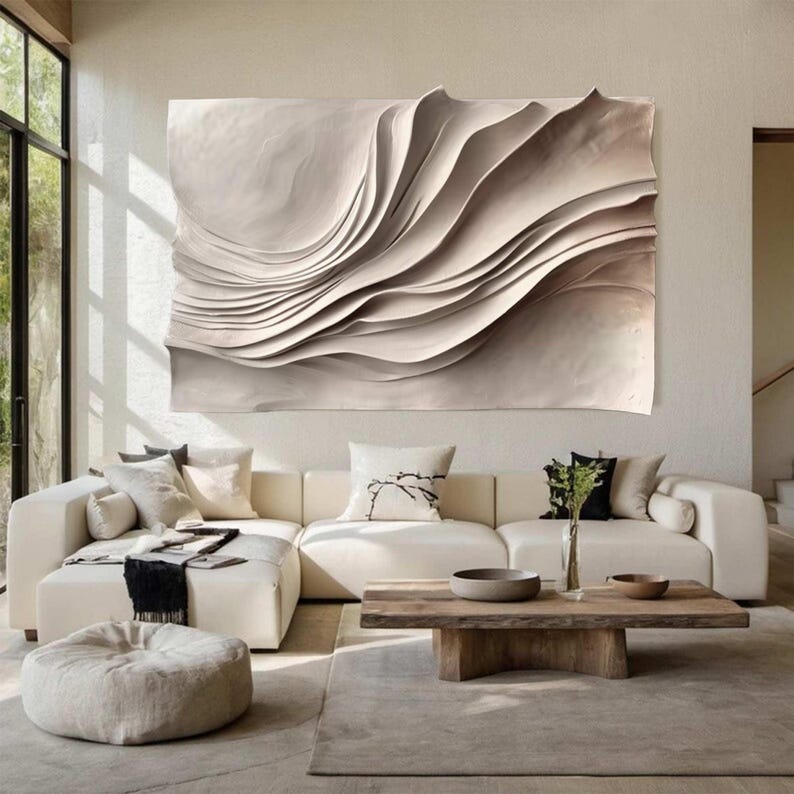 May include: A large, abstract wall art piece with a layered, wave-like design in shades of beige and cream. The artwork hangs above a cream-colored sectional sofa, complementing the neutral tones of the room. A wooden coffee table and a round ottoman complete the living room setting.