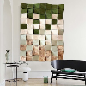 May include: A wall art piece made of wood squares in shades of green, beige, and brown. The squares are arranged in a grid pattern and have a 3D effect. The art piece is hanging on a white wall in a living room setting.