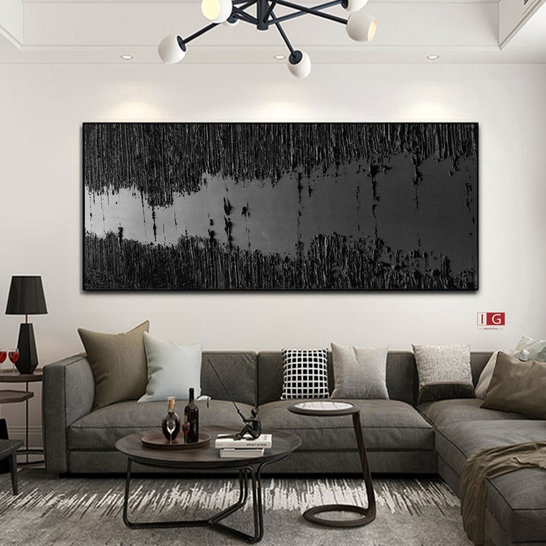 Puede incluir: Una pintura abstracta en blanco y negro con una superficie texturizada. La pintura est&aacute; colgada en una pared blanca sobre un sof&aacute; seccional gris.