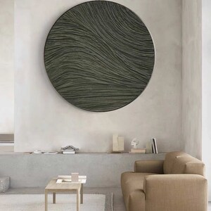 May include: A large, round wall art piece with a textured, wave-like pattern in shades of gray and green. The artwork is framed in black and hangs on a neutral-colored wall. The piece adds a modern, minimalist touch to the room's decor.
