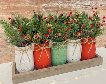 May include: Four mason jars painted in red, white, and green with sprigs of greenery and red berries inside. The jars are arranged in a wooden tray.