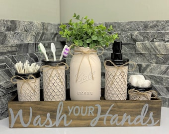 May include: A wooden box with the words "Wash Your Hands" painted on it. The box contains five mason jars, two with toothbrushes, one with cotton swabs, one with a plant, and one with a pump bottle of soap.