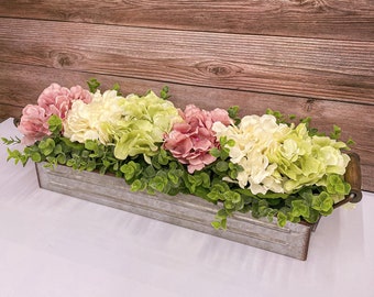 May include: A rectangular metal planter filled with a variety of colorful faux flowers and greenery. The arrangement includes pink, cream, and light green hydrangeas, with green foliage. The planter has a rustic, galvanized metal finish.