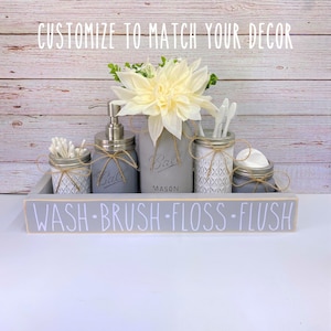 May include: A grey painted wooden tray with the words "WASH-BRUSH-FLOSS-FLUSH" on the front. The tray holds five mason jars, one with a pump bottle of soap, one with cotton buds, one with toothbrushes, one with floss, and one with cotton rounds. The jars are decorated with twine and a white floral arrangement.