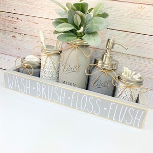 May include: A gray wooden tray with the words "WASH BRUSH FLOSS FLUSH" in white lettering. The tray holds several gray painted mason jars with twine accents, a soap dispenser, and a faux plant arrangement.