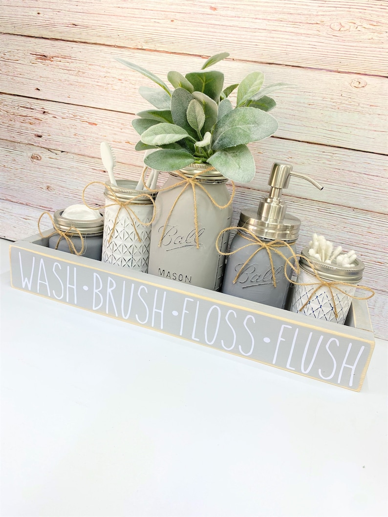 May include: A gray wooden tray with the words "WASH BRUSH FLOSS FLUSH" in white lettering. The tray holds several gray painted mason jars with twine accents, a soap dispenser, and a faux plant arrangement.