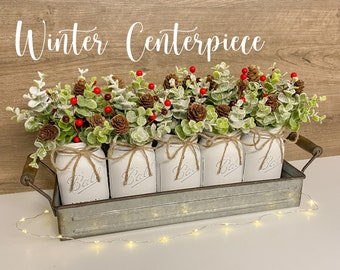 May include: A winter centerpiece featuring five white mason jars filled with greenery, red berries, and pine cones. The jars are arranged in a galvanized metal tray with a rustic wood background. The text "Winter Centerpiece" is displayed above the arrangement.