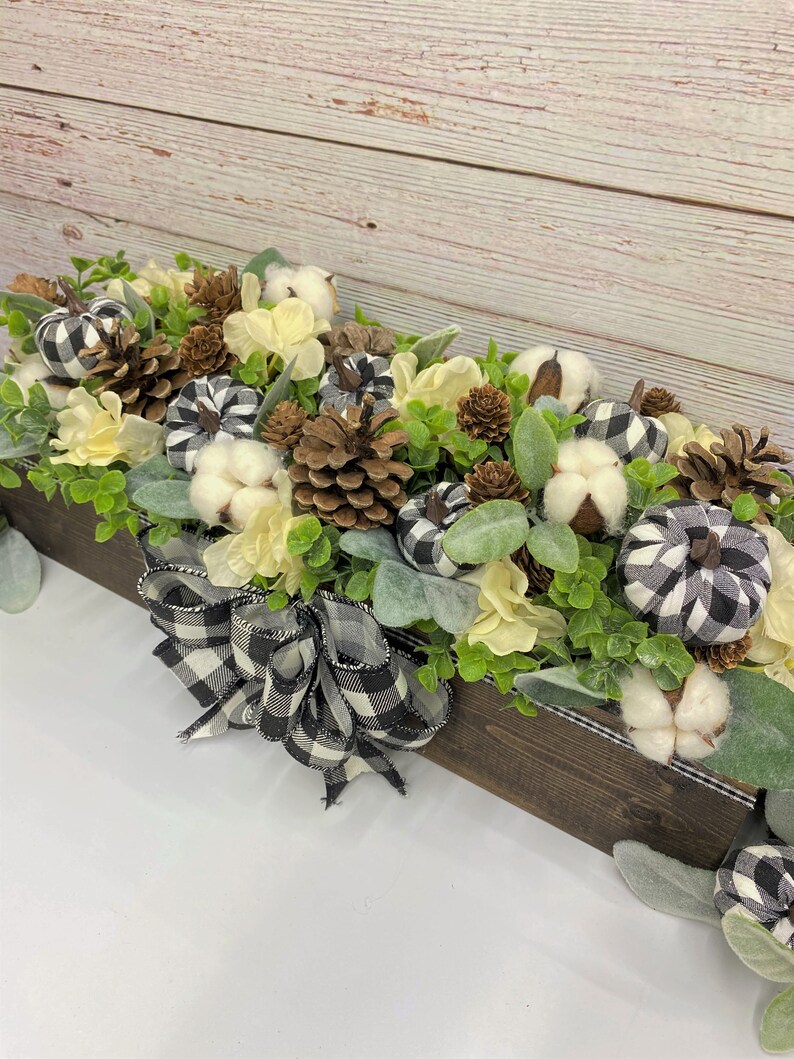 Pu&ograve; includere: Una scatola di legno piena di un arrangiamento autunnale di boccioli di cotone bianco, zucche a scacchi neri e bianchi, pigne e fiori color crema. Un nastro a scacchi neri e bianchi &egrave; legato attorno alla scatola.