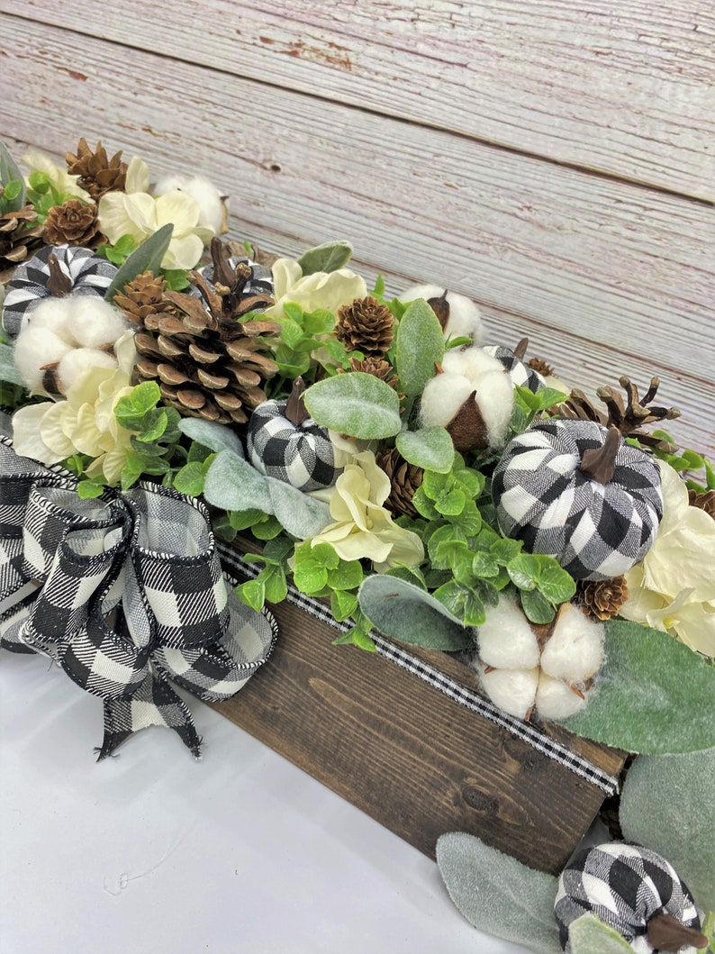 Pu&ograve; includere: Un centrotavola in legno decorato con boccioli di cotone bianco, pigne e zucche in tessuto a scacchi neri e bianchi. La scatola &egrave; decorata con un nastro a scacchi neri e bianchi.