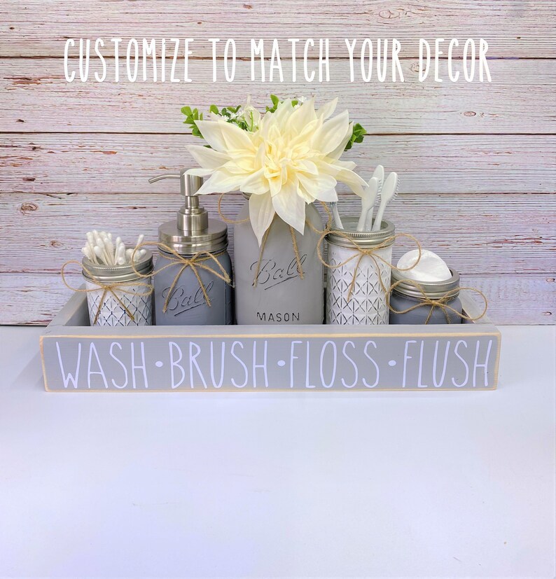 May include: A grey painted wooden tray with the words "WASH-BRUSH-FLOSS-FLUSH" on the front. The tray holds five mason jars, one with a pump bottle of soap, one with cotton buds, one with toothbrushes, one with floss, and one with cotton rounds. The jars are decorated with twine and a white floral arrangement.