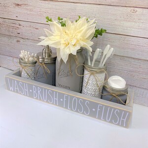 May include: A grey wooden box with the words "WASH - BRUSH - FLOSS - FLUSH" written on it. The box contains five mason jars, four of which are filled with bathroom supplies. The jars are decorated with twine and a white geometric pattern. A white flower arrangement is in the centre of the box.