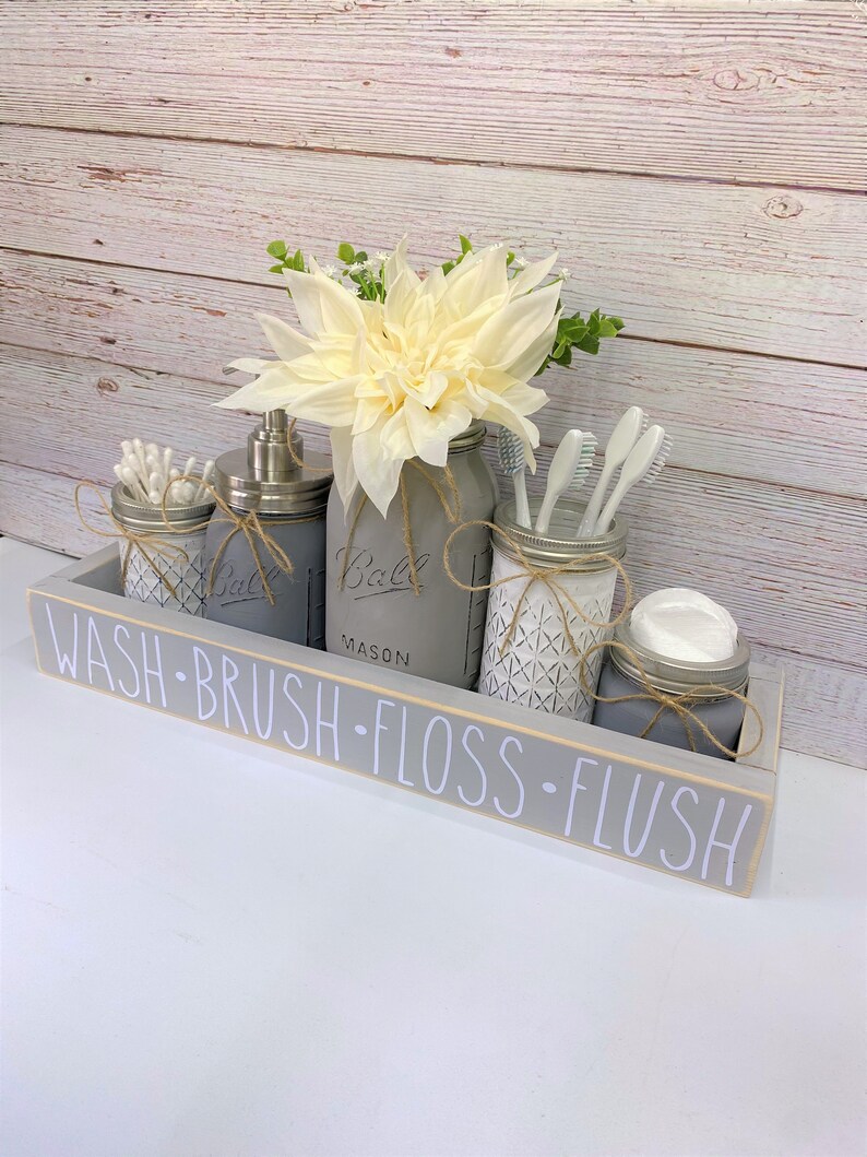 May include: A grey wooden box with the words "WASH - BRUSH - FLOSS - FLUSH" written on it. The box contains five mason jars, four of which are filled with bathroom supplies. The jars are decorated with twine and a white geometric pattern. A white flower arrangement is in the centre of the box.
