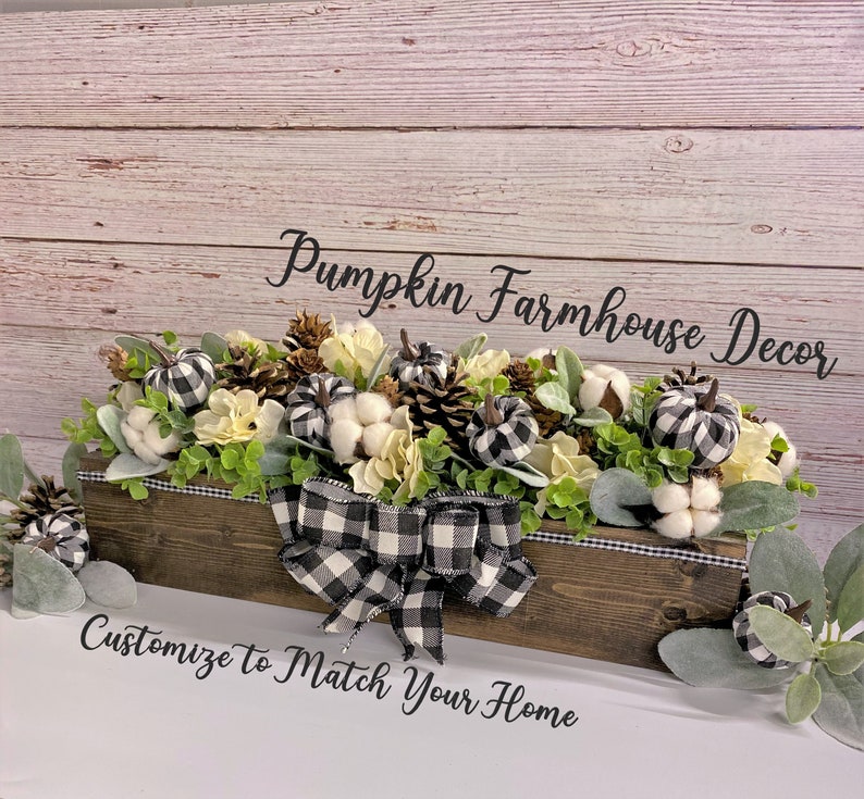 Pu&ograve; includere: Un centrotavola in legno decorato con zucche a scacchi bianchi e neri, boccioli di cotone bianchi, pigne e verde. La scatola &egrave; legata con un nastro a scacchi bianchi e neri e il testo "Pumpkin Farmhouse Decor" e "Customize to Match Your Home" &egrave; visualizzato.