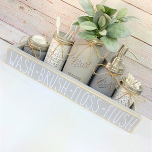 May include: A gray wooden bathroom organizer with the words "WASH - BRUSH - FLOSS - FLUSH" in white. It holds five mason jars, some with lids, and one with a pump. The jars are decorated with twine and contain items like toothbrushes and cotton swabs.