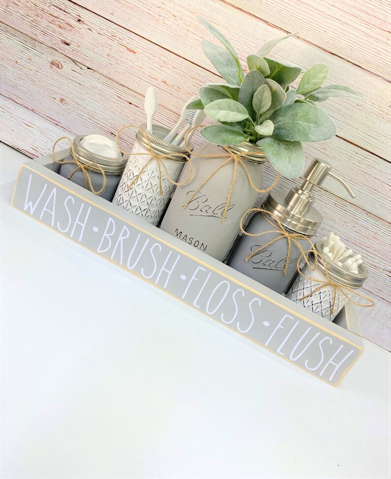 May include: A gray wooden bathroom organizer with the words "WASH - BRUSH - FLOSS - FLUSH" in white. It holds five mason jars, some with lids, and one with a pump. The jars are decorated with twine and contain items like toothbrushes and cotton swabs.