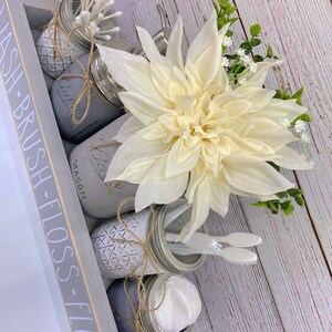 May include: A grey wooden tray with the words "WASH - BRUSH - FLOSS - FLUSH" written on the side. The tray contains four mason jars, each with a different item inside. The jars are decorated with twine and a geometric pattern. The tray also contains a white flower, a small white towel, and two toothbrushes.