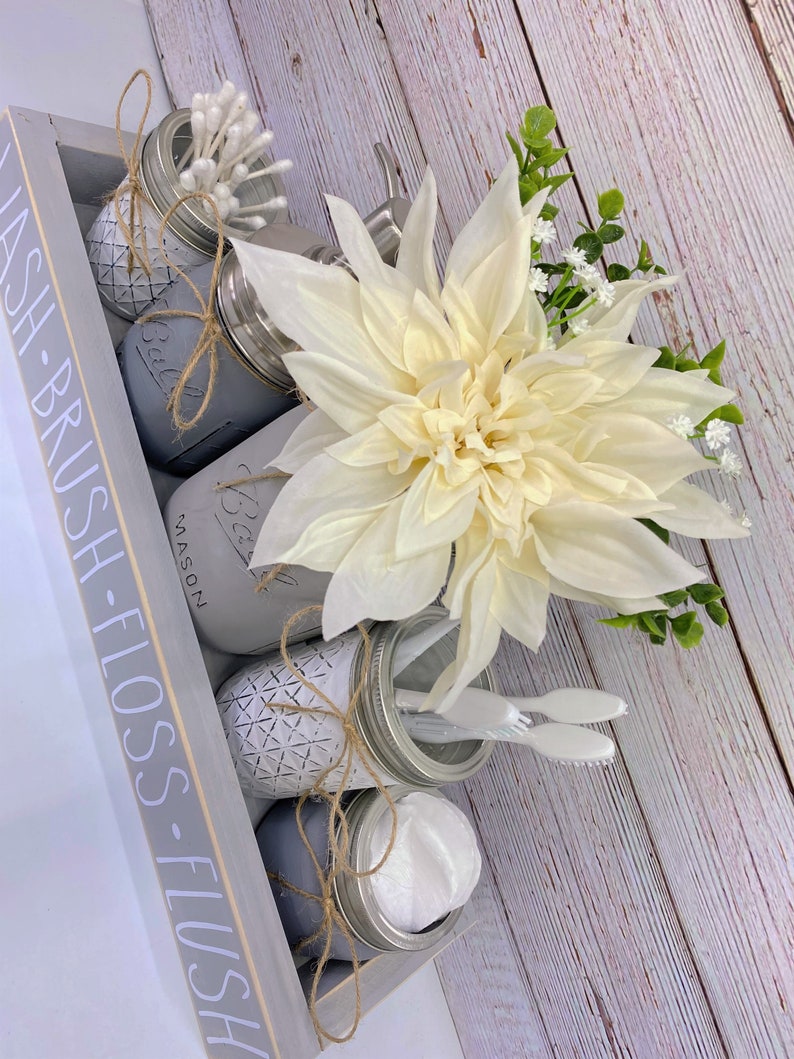 May include: A grey wooden tray with the words "WASH - BRUSH - FLOSS - FLUSH" written on the side. The tray contains four mason jars, each with a different item inside. The jars are decorated with twine and a geometric pattern. The tray also contains a white flower, a small white towel, and two toothbrushes.