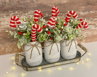 May include: Three white mason jars with red and white candy canes and green sprigs in them. The jars are sitting on a metal tray with a rustic wood background.