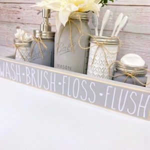 May include: Grey bathroom organiser with the words "Wash - Brush - Floss - Flush" on the front. The organiser has four mason jars with twine and a white flower arrangement. The jars contain cotton buds, toothbrushes, floss, and soap.