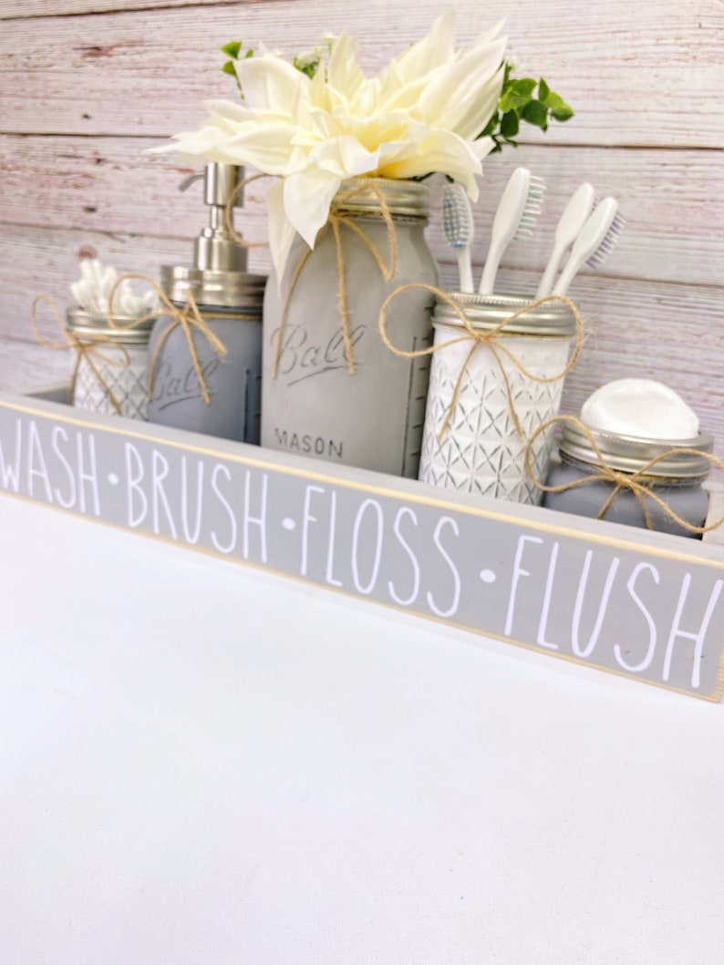 May include: Grey bathroom organiser with the words "Wash - Brush - Floss - Flush" on the front. The organiser has four mason jars with twine and a white flower arrangement. The jars contain cotton buds, toothbrushes, floss, and soap.