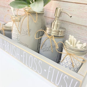 May include: A gray mason jar bathroom set with a wooden tray. The set includes a soap dispenser, jars for cotton swabs, and a vase with greenery. The tray has the words "BRUSH - FLOSS - FLUSH" in white.