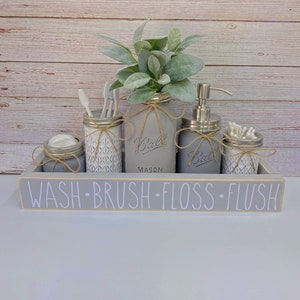 May include: A gray wooden tray with the words "WASH BRUSH FLOSS FLUSH" in white lettering. The tray holds several gray mason jars with twine accents, some with white geometric patterns. The jars contain toiletries and greenery.
