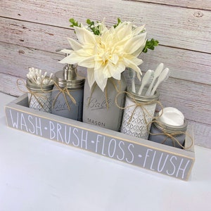 May include: A grey wooden tray with the words "WASH-BRUSH-FLOSS-FLUSH" written on it. The tray holds four mason jars, one with a white flower, one with a pump bottle of soap, one with cotton buds, and one with toothbrushes.