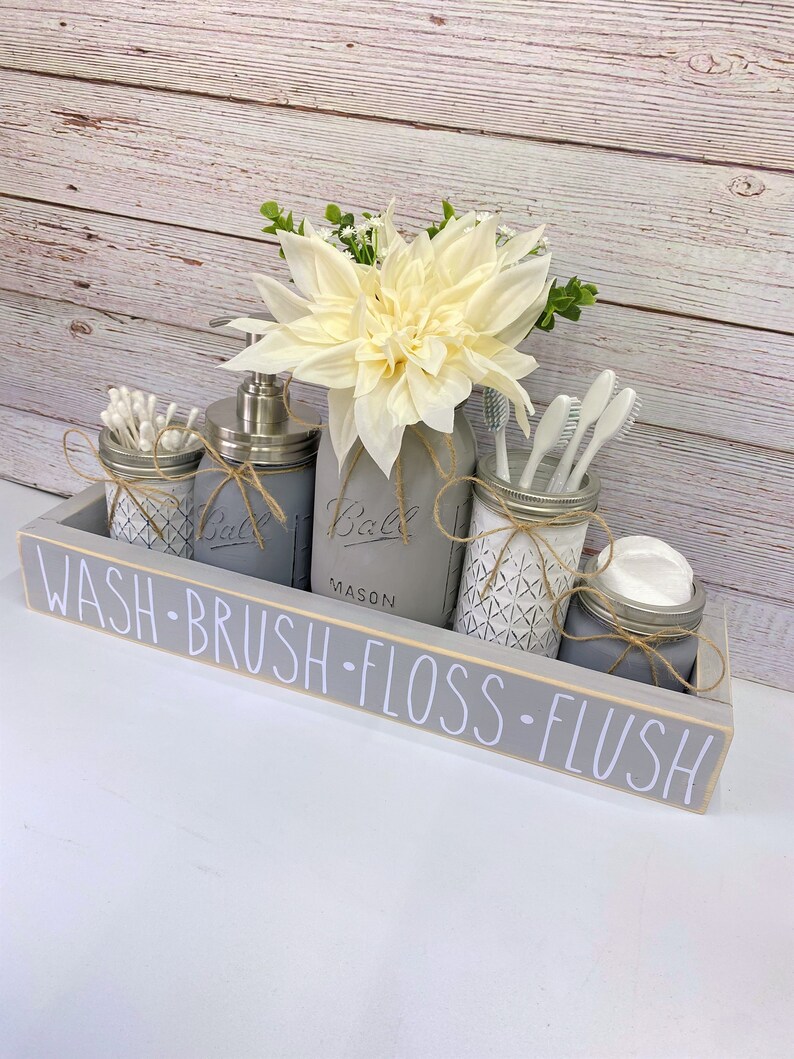 May include: A grey wooden tray with the words "WASH-BRUSH-FLOSS-FLUSH" written on it. The tray holds four mason jars, one with a white flower, one with a pump bottle of soap, one with cotton buds, and one with toothbrushes.