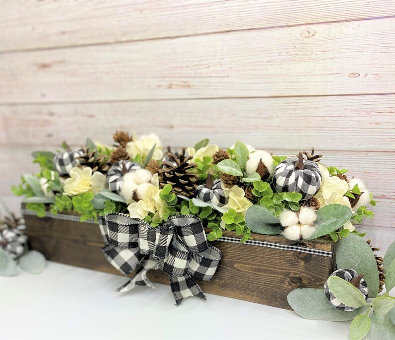Pu&ograve; includere: Un centrotavola in legno decorato con boccioli di cotone bianco, zucche a quadri bianchi e neri, pigne e verde. Un nastro a quadri bianchi e neri &egrave; legato attorno alla scatola.