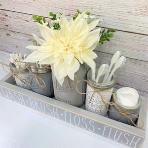 May include: A grey wooden tray with the words "WASH-BRUSH-FLOSS-FLUSH" painted on the front. The tray holds four mason jars, each with a different bathroom item: cotton buds, soap, toothbrushes, and cotton wool balls. A white flower sits in the centre of the tray.