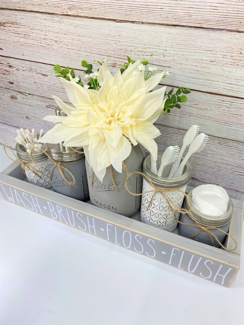 May include: A grey wooden tray with the words "WASH-BRUSH-FLOSS-FLUSH" painted on the front. The tray holds four mason jars, each with a different bathroom item: cotton buds, soap, toothbrushes, and cotton wool balls. A white flower sits in the centre of the tray.