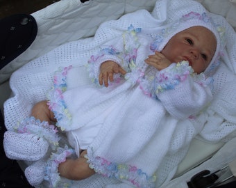 May include: A white knitted baby doll outfit with a ruffled trim and a matching bonnet. The doll is lying in a white crib.