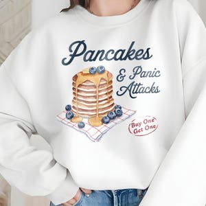 May include: White sweatshirt with a graphic of a stack of pancakes with blueberries and syrup, and the text "Pancakes & Panic Attacks" and "Buy One Get One".