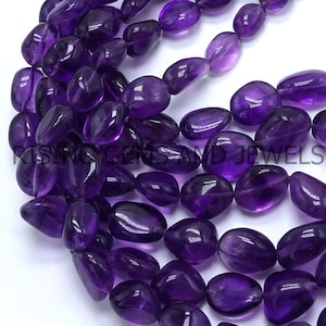 May include: Close-up of strands of polished, deep purple amethyst gemstone beads. The beads are oval-shaped and have a glossy finish. The image shows multiple strands, with the text "RISING GEMS AND JEWELS" partially visible.