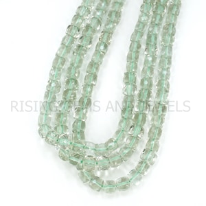 May include: Several strands of light green, square-cut gemstone beads. The beads are strung on a light green cord. The image includes the text "RISING GEMS AND JEWELS" in the lower left corner.