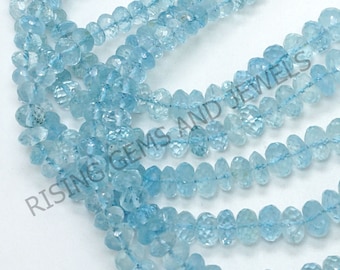 May include: A strand of light blue aquamarine beads, faceted and with a slightly irregular shape. The beads are strung on a clear thread and are approximately 6mm in diameter. The beads are arranged in a loose, flowing pattern. The image is taken from a close-up perspective, showing the detail of the beads. The text "RISING GEMS AND JEWELS" is visible in the image.
