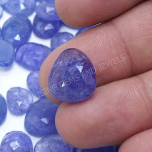 Peut inclure: Une pierre précieuse bleue en forme de goutte d'eau, facettée, est tenue entre les doigts, avec d'autres pierres similaires en arrière-plan. La gemme est translucide, avec des facettes internes visibles. Le texte "RISING GEMS AND JEWELS" est partiellement visible.