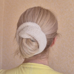 May include: A knitted white hair accessory, possibly a bun cover, encircles a blonde bun. The person is wearing a yellow top. The background is a light beige colour.