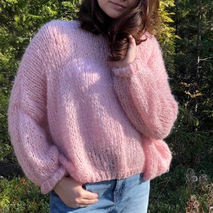 Peut inclure: Un pull rose clair en tricot surdimensionné avec de longues manches volumineuses. Le pull a un col rond et une coupe légèrement courte. Le modèle porte un jean bleu. L'arrière-plan est un cadre extérieur naturel avec un feuillage vert.