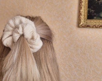 May include: A light beige knitted scrunchie holds back long, light brown hair. The scrunchie is made of a thick, textured yarn. The hair is styled in a ponytail. The background is a light beige wall with a framed picture.
