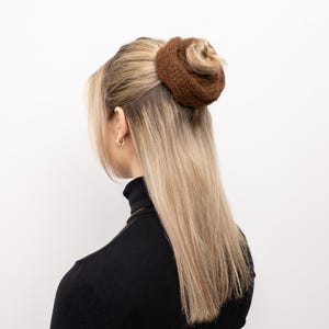 Op de afbeelding: Een bruin gebreid haaraccessoire in de vorm van een knot. Het haaraccessoire wordt op de achterkant van het hoofd gedragen, met lang blond haar. De persoon draagt een zwarte coltrui.