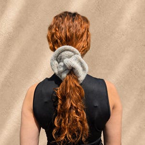 May include: A light gray, chunky knit hair scrunchie with a soft, fuzzy texture. The accessory is gathered in a circular shape, featuring visible yarn loops and a thick, voluminous construction that secures a section of long, wavy, auburn hair.