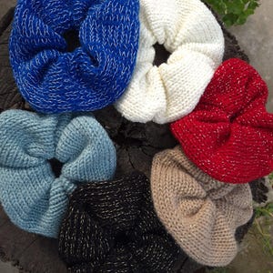 May include: A collection of six knitted scrunchies in various colors, including blue, white, red, light blue, black, and tan. Each scrunchie has a textured appearance with a subtle metallic thread woven throughout.