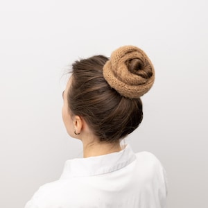 Può includere: I capelli di una donna sono acconciati a chignon, con un accessorio per capelli lavorato a maglia. L'accessorio per capelli è di colore marrone chiaro ed è avvolto attorno allo chignon. La donna indossa una camicia bianca con colletto. Lo sfondo è bianco.