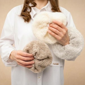 May include: Three knitted berets in cream, light grey, and tan are held in front of a person wearing a white shirt. The berets are made of a chunky knit material, and the colours are neutral and versatile.