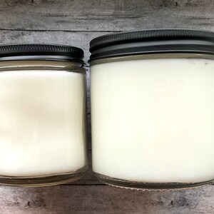May include: Two glass candle jars with black lids. The jars are filled with white wax and are sitting on a wooden surface.